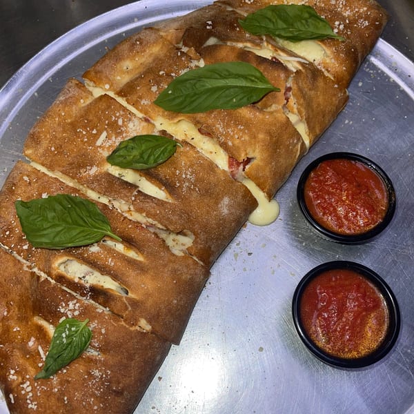 A golden-brown calzone, sliced open to reveal melted cheese and fillings, garnished with basil and served with two cups of marinara sauce.