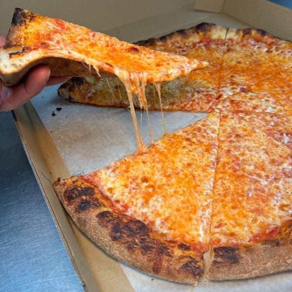 Hand lifting a cheesy slice of Napoli Pizza from a whole pie in a cardboard box.
